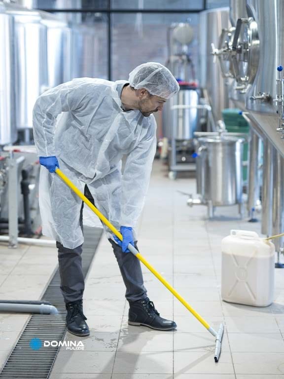 Un operaio in tuta protettiva bianca, retina per capelli e guanti blu pulisce il pavimento con uno spazzolone in un impianto industriale sanitario con vasche in acciaio inox, dimostrando il livello di professionalità tipico di Pulizie Industriali.