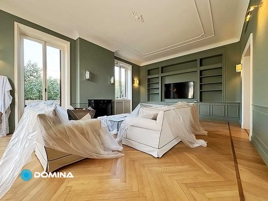 Un ampio soggiorno con pareti verdi, mobili ricoperti da teli protettivi da un'impresa di pulizie, pavimento in legno a spina di pesce, ampie finestre e uno scaffale a muro con TV montata.