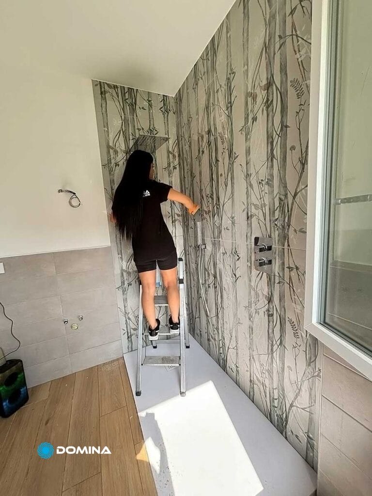 Una persona è in piedi su una scala, mentre applica carta da parati con un motivo naturalistico a una parete in una stanza parzialmente piastrellata. Domina Pulizie potrebbe fornire un eccezionale servizio di pulizie di fine cantiere a Desio per garantire che lo spazio rimanga incontaminato durante e dopo i progetti di ristrutturazione della casa.