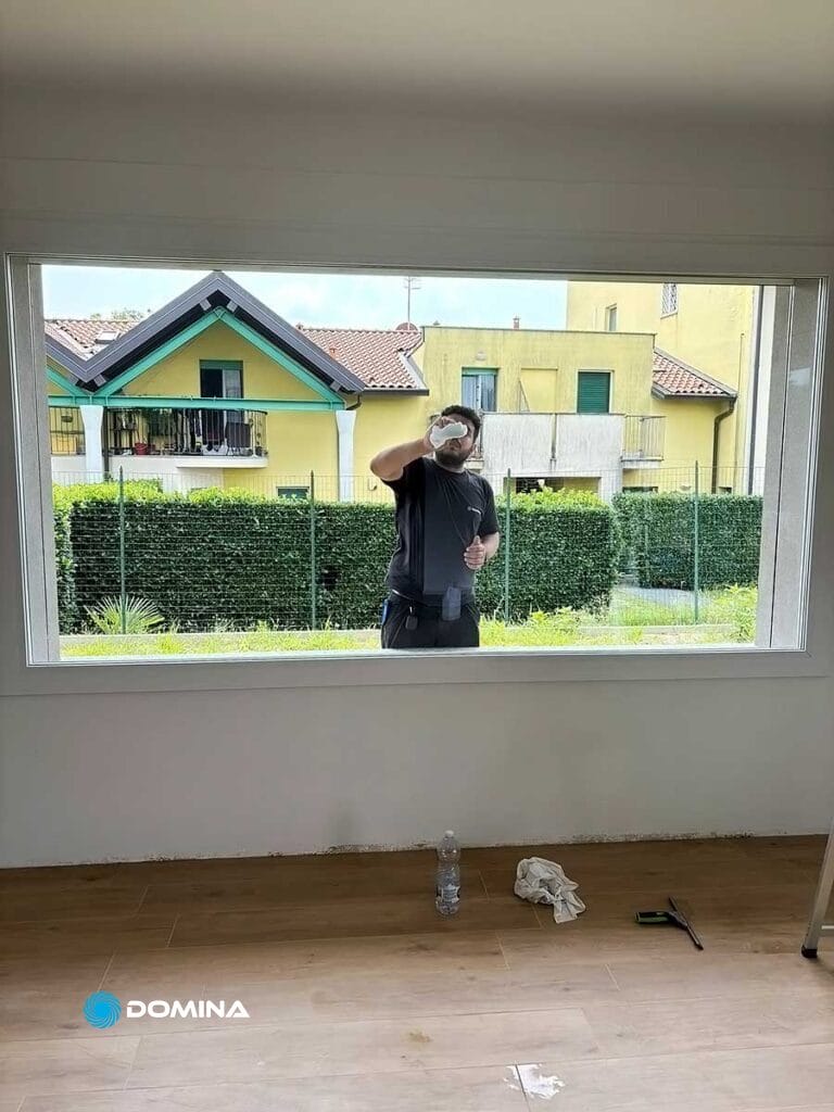 Una persona di Domina Pulizie pulisce l'esterno di una grande finestra in una stanza con pavimento in legno. Sul pavimento accanto a loro ci sono una bottiglia di soluzione detergente, uno straccio e un cacciavite. Fuori si vedono case e vegetazione. Fa parte del loro completo servizio di pulizie di fine cantiere a Desio.