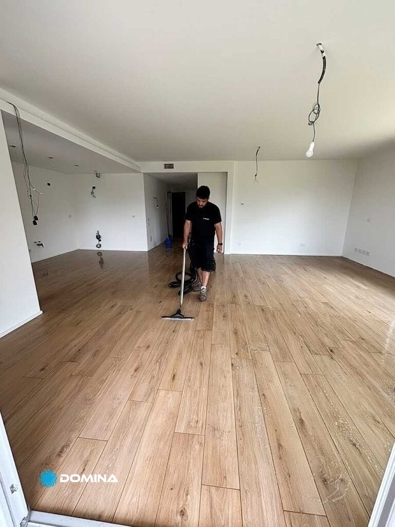 Una persona di Domina Pulizie passa l'aspirapolvere in una spaziosa stanza con pavimento in legno. La stanza ha pareti bianche e cavi a vista che pendono dal soffitto e dalle pareti, come parte del loro servizio di pulizie di fine cantiere a Desio.