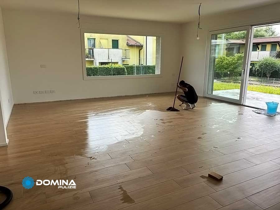 Una persona lava il pavimento in legno appena installato in una stanza vuota e luminosa con grandi finestre e una porta scorrevole in vetro. I prodotti per la pulizia del servizio di pulizie di fine cantiere a Desio di Domina Pulizie sono visibili sul pavimento. All'esterno si possono vedere edifici e vegetazione.