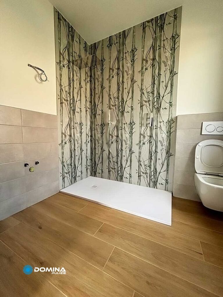 Un bagno con cabina doccia con carta da parati a tema arboreo, pareti piastrellate beige, pavimento in legno e water bianco. Recentemente revisionato dal servizio di pulizie di fine cantiere di Domina Pulizie a Desio.