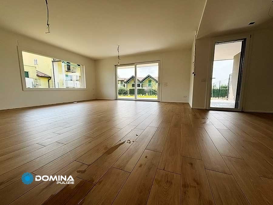 Stanza vuota con pavimento in legno, grandi finestre e porte che conducono a un'area esterna; fili che pendono dal soffitto e visibili segni di sporco sul pavimento. Contatta Domina Pulizie per un servizio professionale di pulizie di fine cantiere a Desio per riportare lo spazio alle sue condizioni originali.