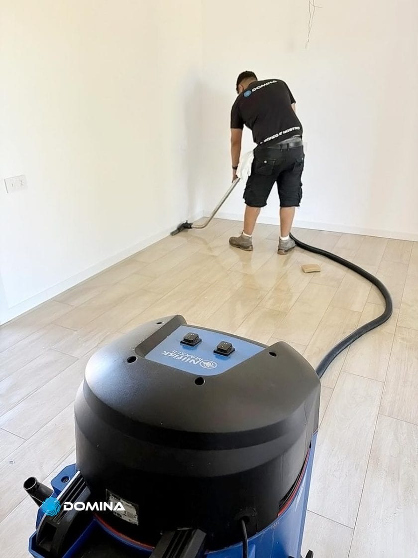 Persona che usa un aspirapolvere per pulire un pavimento in legno chiaro in una stanza con pareti bianche.