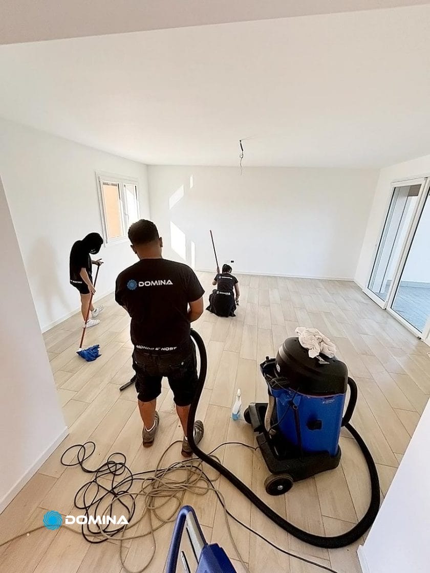 Due persone puliscono una stanza vuota con un aspirapolvere e uno straccio. Un aspirapolvere è in primo piano. La stanza ha pavimenti in legno chiaro e pareti bianche.