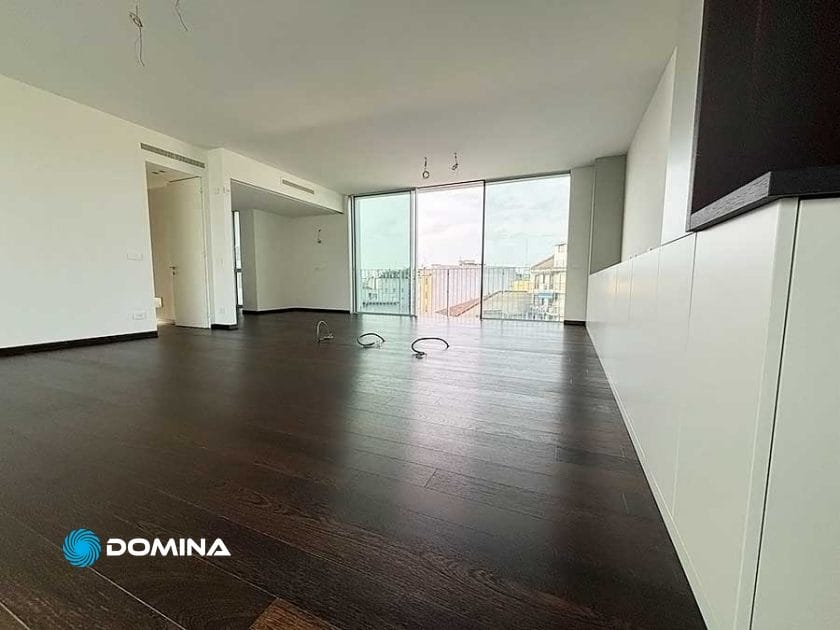 Un moderno appartamento non arredato a Milano con pavimento in legno scuro, pareti bianche, ampie finestre dal pavimento al soffitto e vista sul balcone. Perfetto per chi cerca servizi professionali e pulizie di lusso per appartamenti.