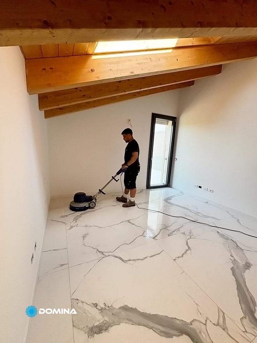 Una persona sta lucidando meticolosamente grandi piastrelle di marmo in un'elegante stanza con travi in legno sul soffitto e un lucernario, dando vita all'essenza di un Attico. Questa scena si svolge a Buccinasco, dove è in mostra l'esperta Pulizie Fine Cantiere.