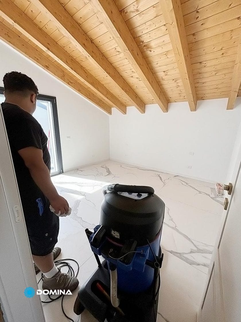 In una stanza luminosa con travi in legno e pavimento effetto marmo, una persona in un attico aziona un aspirapolvere blu, occupandosi meticolosamente delle pulizie di fine cantiere.