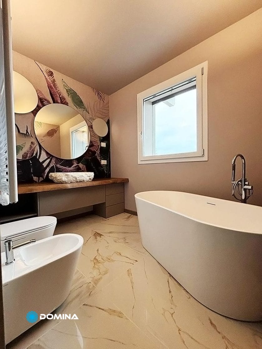 Il bagno moderno nel Centro di Milano è dotato di una vasca da bagno indipendente, un bidet e un grande specchio su cui è appoggiata una carta da parati con motivi a foglie. Una finestra lascia entrare la luce naturale sulla finitura marmorea del pavimento, catturando perfettamente l'essenza di rinnovare il lusso nella vita urbana.