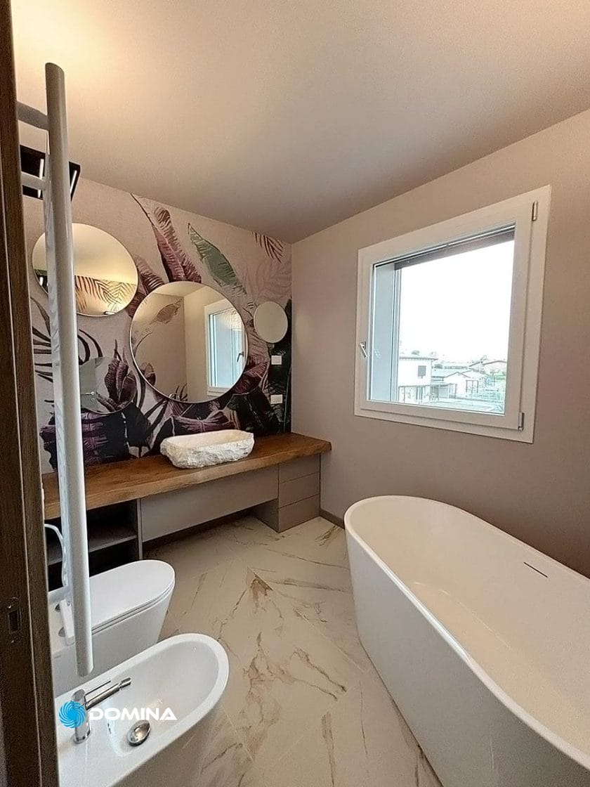 Bagno moderno con vasca bianca, bidet e water. Piano di lavoro in legno con lavandino a catinella e numerosi specchi rotondi. Ampia finestra con vista sullo skyline del centro di Milano, mentre la parete con motivo floreale aggiunge fascino. Perfetto per chi desidera rinnovare il lusso a casa.