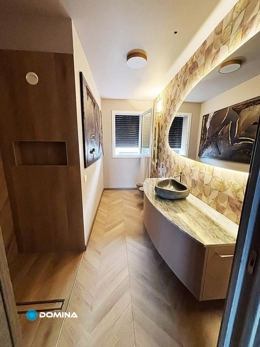 Un bagno moderno nel cuore del Centro di Milano, con un grande piano di lavoro in marmo curvo e un lavandino a catinella. Il pavimento in legno a spina di pesce e le piastrelle geometriche delle pareti trasudano eleganza, completate da due opere d'arte incorniciate e una finestra con persiane chiuse, perfette per chi cerca di rinnovare il lusso.