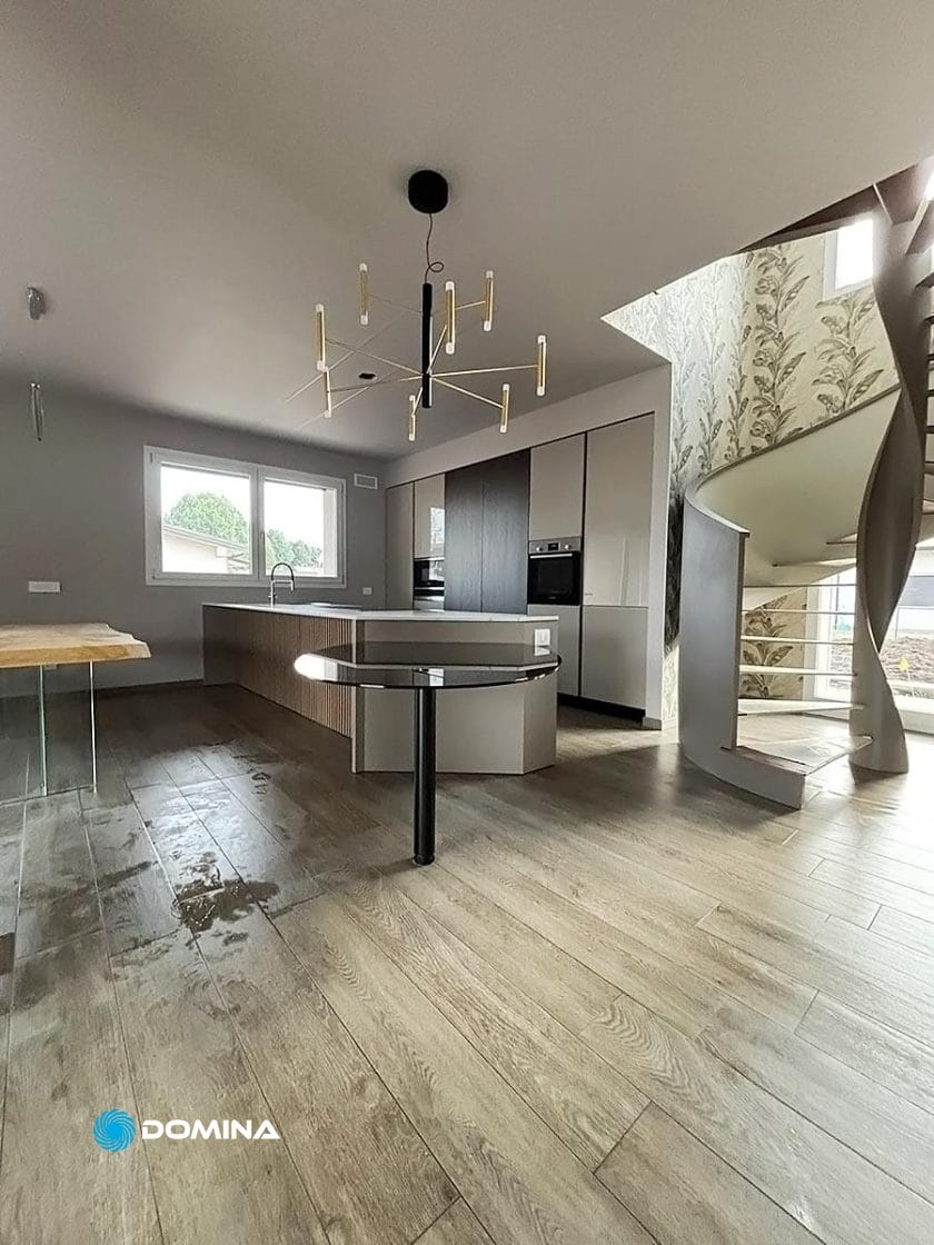 Cucina moderna con pavimento in legno, bancone a isola e una scala a chiocciola unica. Una finestra fornisce luce naturale e un lampadario pende dal soffitto. Ideale per chi cerca pulizie di fondo appartamento di lusso nel centro di Milano per mantenere le sue condizioni immacolate.