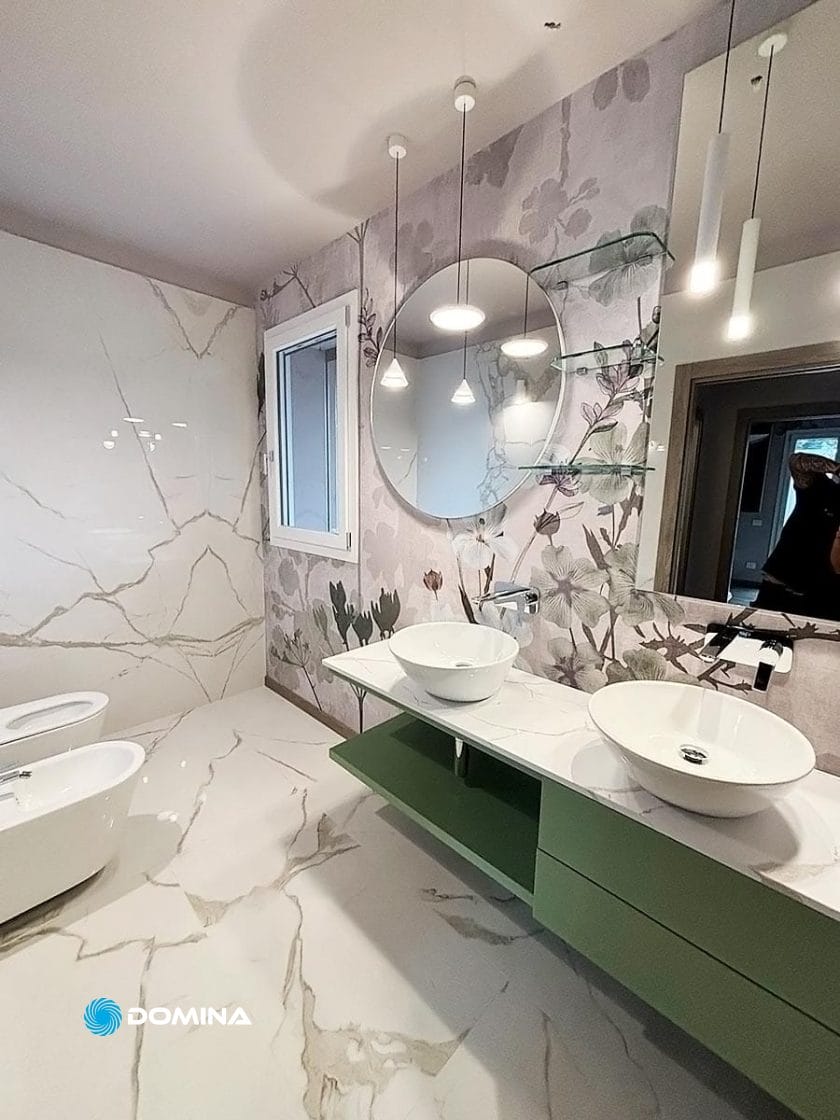 Questo moderno bagno nel cuore del Centro di Milano è caratterizzato da un grande specchio rotondo, doppi lavandini su un mobiletto verde, carta da parati floreale, eleganti luci pendenti e un water con bidet contro pareti e pavimenti in marmo di lusso. Rinnovare il Lusso è evidente in ogni dettaglio.