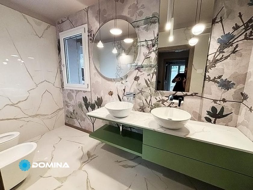 Scoprite un bagno moderno nel cuore del centro di Milano, con carta da parati floreale, due lavabi a catinella su un piano in marmo e un tocco di eleganza con specchi rotondi e lampade a sospensione: una vera testimonianza di Rinnovare il Lusso.
