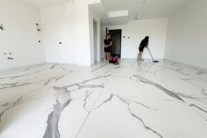 Due persone di Domina Pulizie sono impegnate a pulire una stanza a Milano, caratterizzata da grandi piastrelle con motivi in marmo bianco e pareti bianche immacolate. Una sta lavando il pavimento, assicurandosi un trattamento pavimenti impeccabile, mentre l'altra è in piedi vicino alla porta.