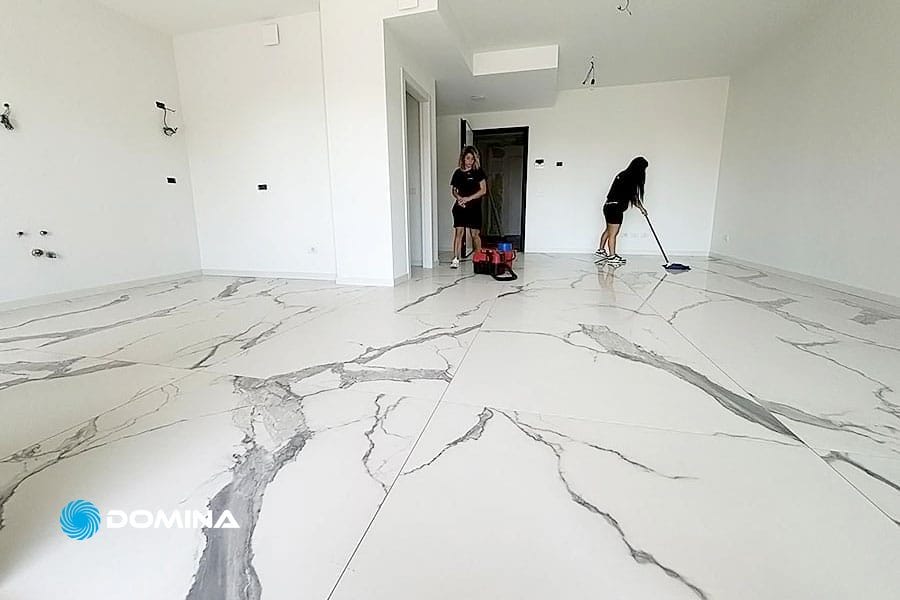Due persone di Domina Pulizie sono impegnate a pulire una stanza a Milano, caratterizzata da grandi piastrelle con motivi in marmo bianco e pareti bianche immacolate. Una sta lavando il pavimento, assicurandosi un trattamento pavimenti impeccabile, mentre l'altra è in piedi vicino alla porta.