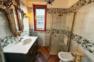 Un bagno con piastrelle a motivi floreali è dotato di una doccia in vetro, un lavandino con uno specchio decorativo e un bidet sotto una finestra con le sbarre, il tutto tenuto in maniera impeccabile, come se fosse stato realizzato da un'impresa di pulizie di Como.