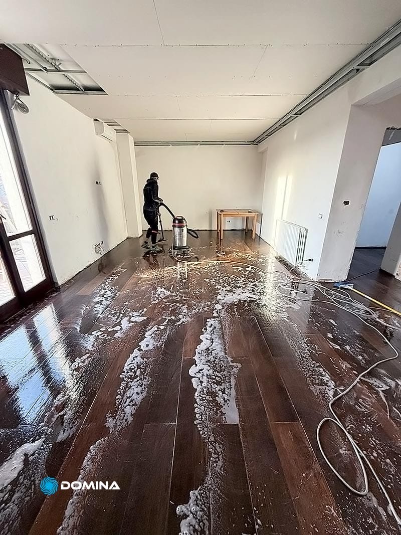 Una persona pulisce meticolosamente una grande stanza dopo la ristrutturazione, usando un aspirapolvere e sapone sul pavimento di legno scuro. Nell'angolo c'è un tavolo, mentre un tubo flessibile è lì vicino. Le pareti bianche e il soffitto parzialmente aperto contribuiscono alla trasformazione mentre tutto inizia a tornare come nuovi.