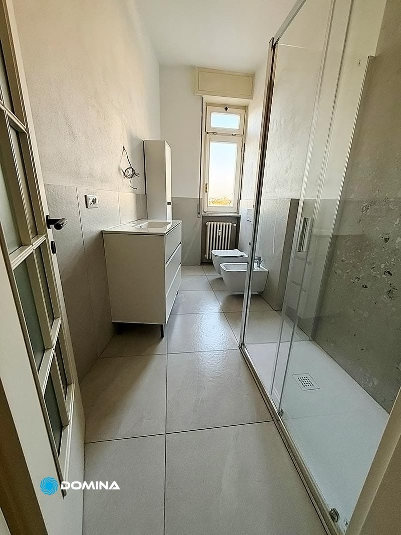 Bagno stretto con lavandino e mobiletto a sinistra, water e bidet sul retro e una doccia con cabina in vetro a destra. Una piccola finestra sopra un termosifone si trova in fondo alla stanza. Questa accogliente casa vanta servizzi professionali per tutte le tue esigenze di pulizia.