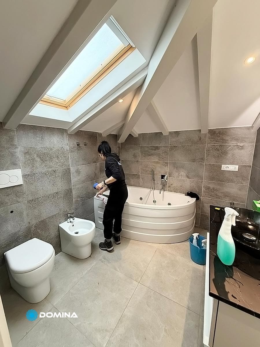 Una persona che esegue meticolosamente pulizie straordinarie in un bagno moderno con lucernario, vasca idromassaggio, water e bidet a Castronno. I prodotti per la pulizia sono disposti ordinatamente sul pavimento e sul bancone, riflettendo l'estetica post-ristrutturazione della villa.