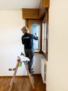 All'interno di una villa post-ristrutturazione, una persona è in piedi su una scala e pulisce meticolosamente una finestra. L'elegante pavimento in legno riflette la cura e la precisione delle pulizie straordinarie in questo spazio splendidamente restaurato.