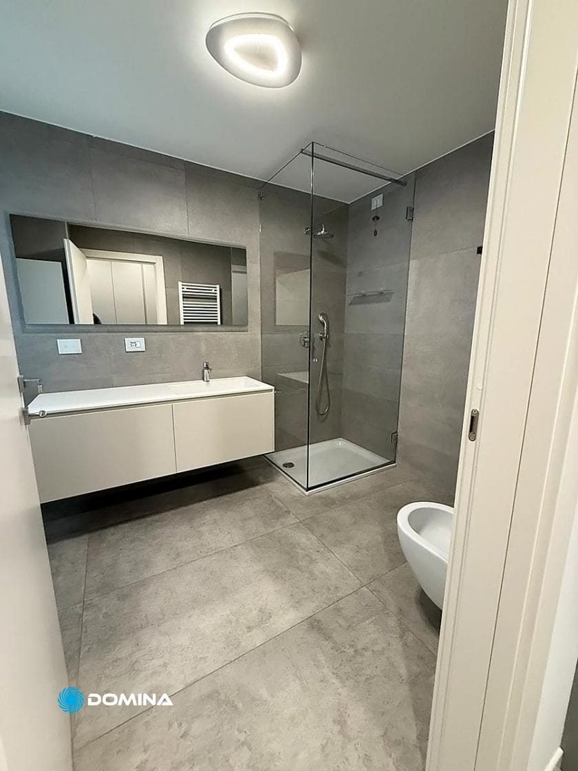 Un bagno moderno con doccia in vetro, lavandino a parete, specchio e WC su un pavimento piastrellato sotto luci a soffitto, splendente come se fosse appena stato pulito.