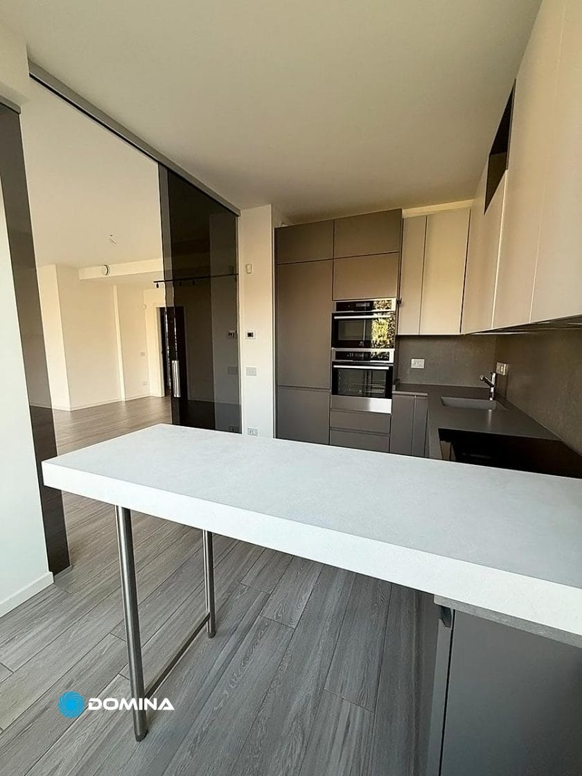 Cucina moderna con mobili grigi eleganti, elettrodomestici in acciaio inox, piano di lavoro bianco e pavimento in legno. Le porte scorrevoli in vetro conducono a una zona soggiorno aperta, perfetta per chi cerca pulizie di lusso a City Life Milano.