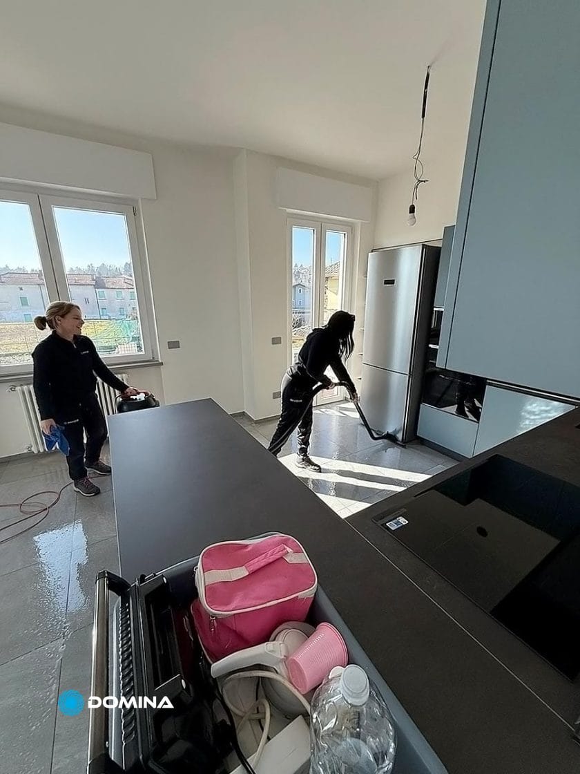 In una cucina moderna ed elegante con ampie finestre, due persone mettono in mostra la loro professionalità. Una passa l'aspirapolvere sul pavimento con perizia, mentre l'altra tiene in mano i prodotti per la pulizia, pronti per pulizie fine cantiere. Le superfici brillano sotto la luce naturale di Trezzano sul Naviglio.
