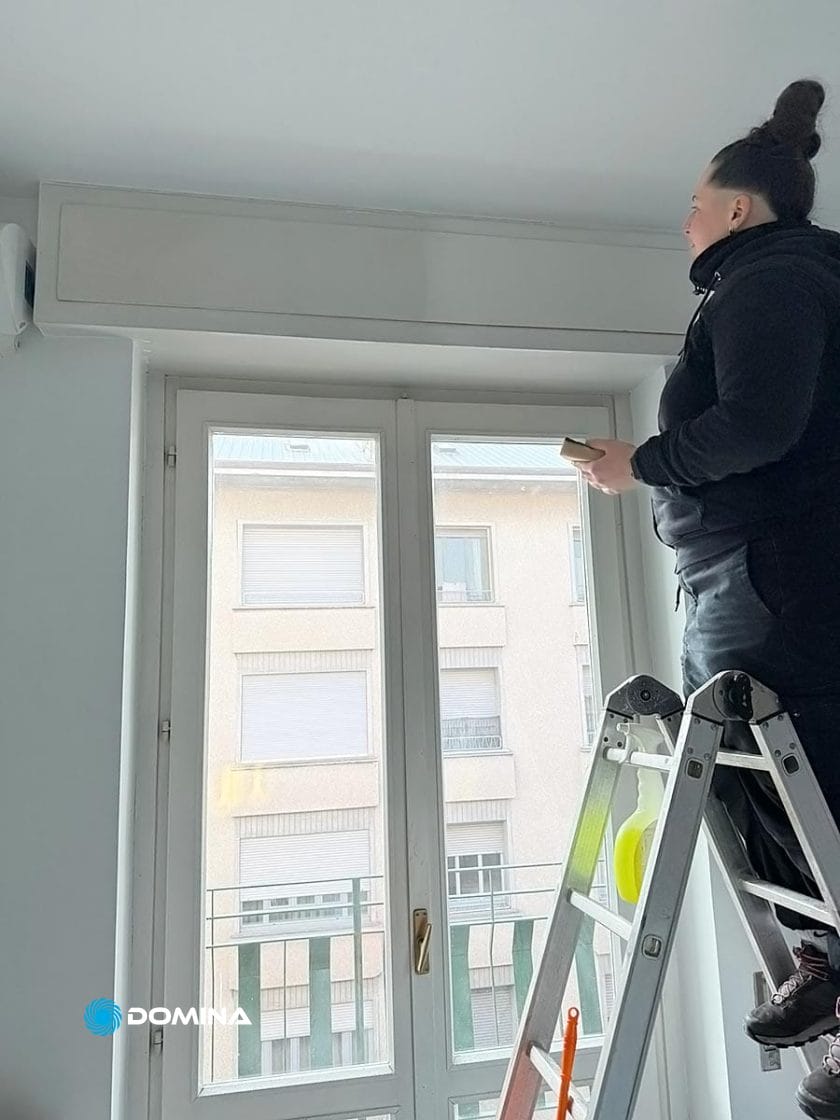 Una persona è in piedi su una scala e ispeziona professionalmente il telaio di una finestra in una stanza molto luminosa, che ricorda un elegante appartamento a Cardano al Campo, con vista sull'edificio esterno.
