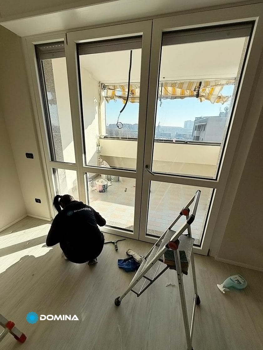 Inginocchiata vicino a una grande porta a vetri in una stanza con pavimento in legno chiaro, una persona dimostra professionalità mentre pulisce. Una scala a pioli e prodotti per la pulizia sono lì vicino, a dimostrazione di una pulizia appartamento meticolosa. Un balcone con vista sulla città si estende verso Cardano al Campo all'esterno.
