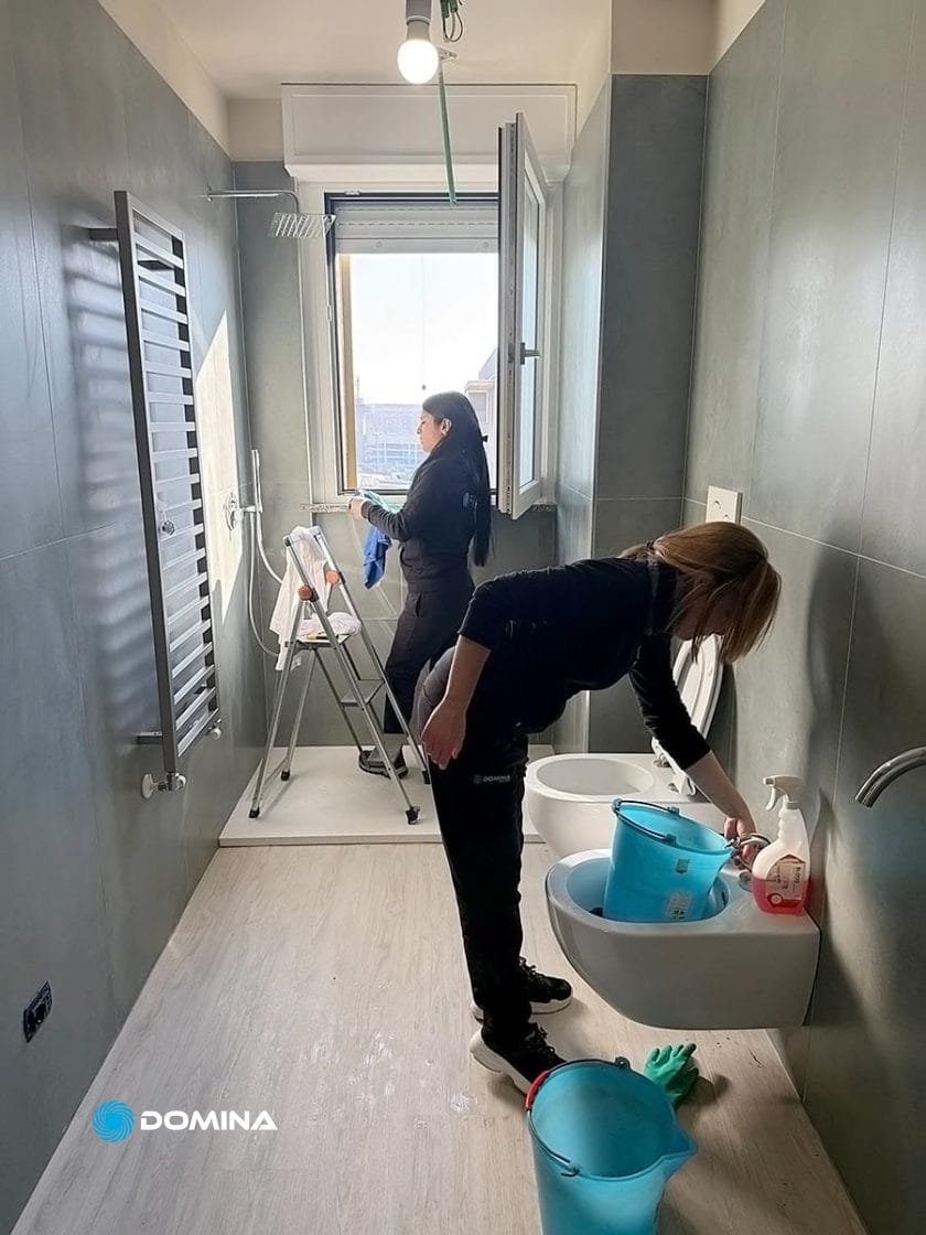 Due persone stanno pulendo diligentemente un bagno. Una è in piedi su una scala a pioli, pulendo una finestra, mentre l'altra lucida il lavandino. Prodotti per la pulizia e secchi sono sul pavimento, assicurando che i loro sforzi raggiungano risultati garantiti.
