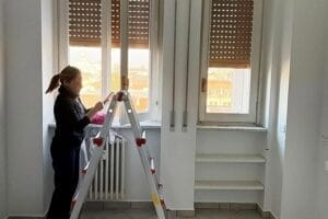 Una persona è in piedi su una scala accanto a una finestra, mentre sistema le tapparelle in una stanza luminosa con pavimento piastrellato. L'ambiente emana un'aura di professionalità, che ricorda i servizi di pulizia appartamento di esperti a Cardano al Campo.