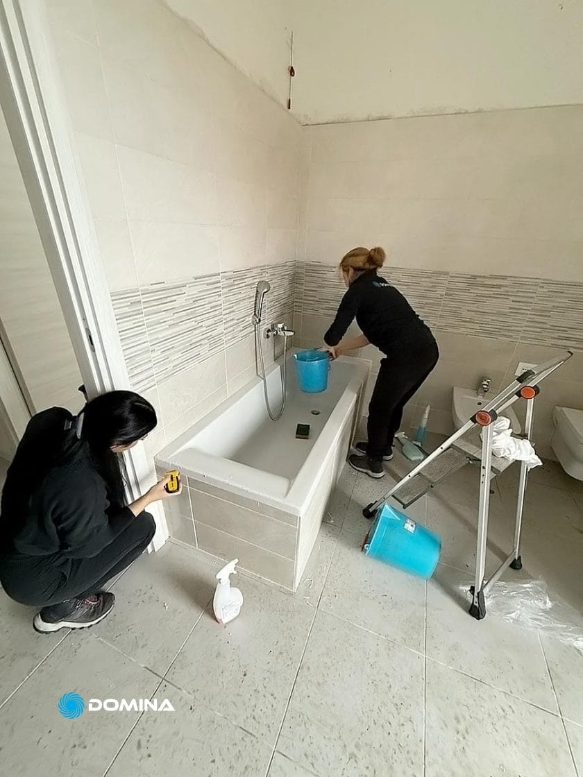 Due persone stanno pulendo diligentemente un bagno, preparandosi per un'ispezione di locazione di fine locazione. Una è vicino alla vasca da bagno sulla destra, mentre l'altra è accovacciata vicino a un muro sulla sinistra. Gli strumenti per la pulizia sono sparsi sul pavimento di questa casa di Busto Arsizio.