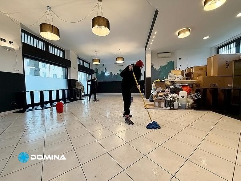 Una persona sta lavando il pavimento in una stanza piena di scatole di cartone e prodotti per la pulizia, incarnando l'essenza di un Servizio Professionale. Sullo sfondo, un altro membro del team di un'Impresa di Pulizie si sta preparando per i suoi compiti.