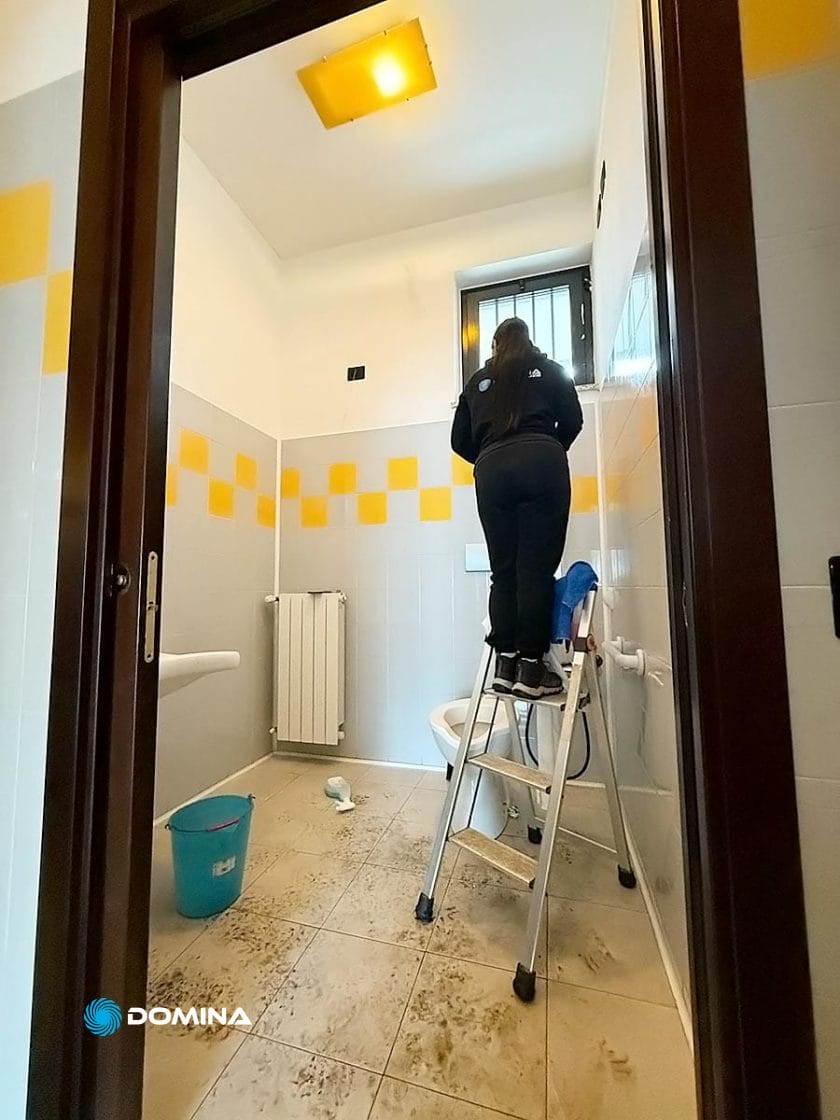 Una persona è in piedi su una scala mentre ripara una finestra in un bagno con piastrelle gialle e bianche. Un secchio per il mocio e diversi articoli per la pulizia, forniti da Impresa di Pulizie, sono sparsi sul pavimento, per garantire che tutto rimanga immacolato dopo le riparazioni.