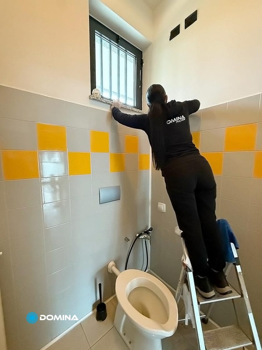 In un bagno di Milano, una persona è in piedi su una scala, mentre sistema con perizia una tenda da finestra sopra il water. Le pareti piastrellate grigie e arancioni aggiungono un tocco moderno a questa ambientazione.