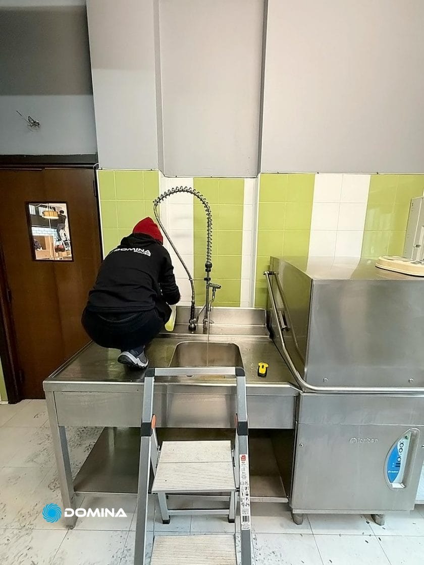 Una persona in giacca nera è inginocchiata su un lavandino in acciaio inox, mentre pulisce il paraschizzi con una precisione professionale che ricorda gli standard meticolosi del Ristorante Milano. Un rubinetto alto e una scala a pioli sono in primo piano, a ricordare l'efficienza di un'Impresa di Pulizie.
