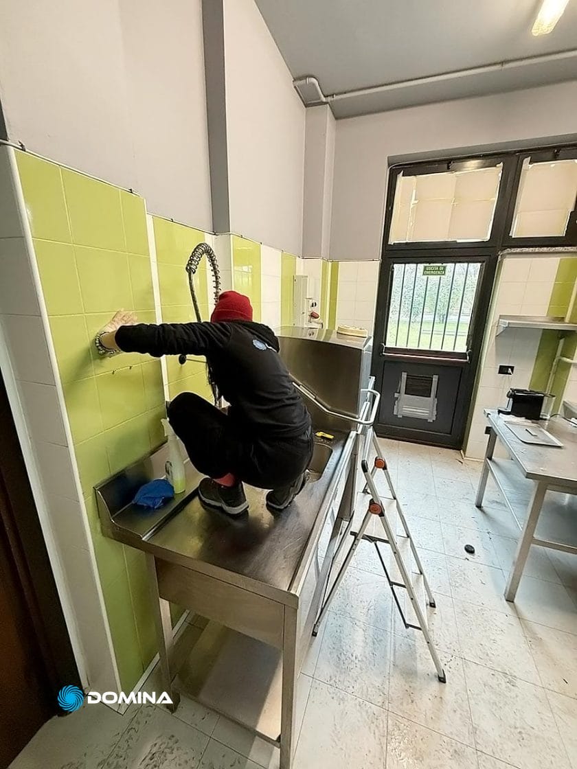 Un addetto di Domina Pulizie è accovacciato su un bancone in acciaio inox e lavora diligentemente a un elemento decorativo a parete nella sala piastrellata del Ristorante Milano, con una scala nelle vicinanze per un facile accesso.