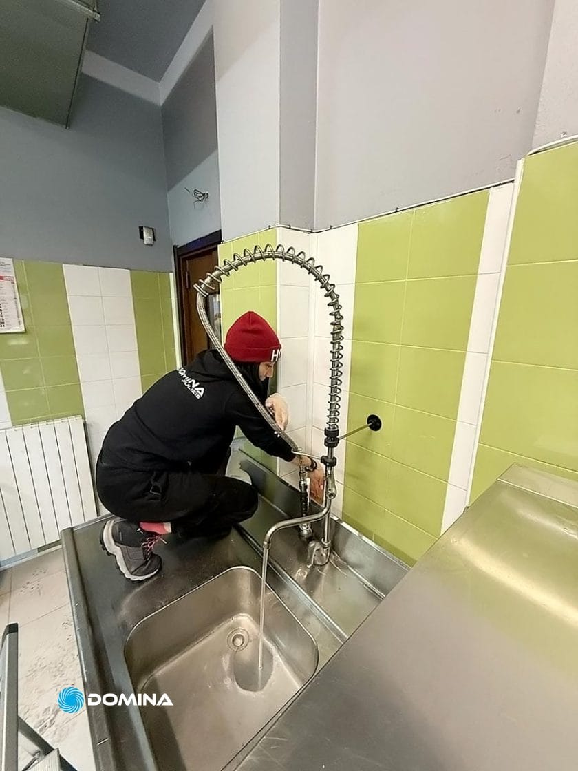 Una persona con un cappello rosso e abiti scuri è accovacciata sul tavolo del lavello della cucina, mentre regola con perizia il flessibile rubinetto commerciale contro una parete con piastrelle verdi e bianche, dimostrando un vero Servizio Professionale simile agli standard del Ristorante Milano.