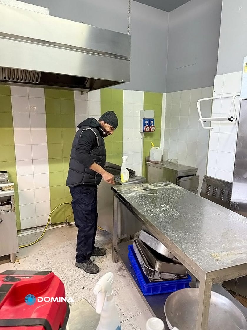 Una persona di un Servizio Professionale pulisce una superficie della cucina con spray e panno. L'area vanta elettrodomestici in acciaio inossidabile, pareti piastrellate di verde e bianco e una cappa di ventilazione, una scena che potrebbe essere tratta direttamente da un Ristorante Milano Centro.