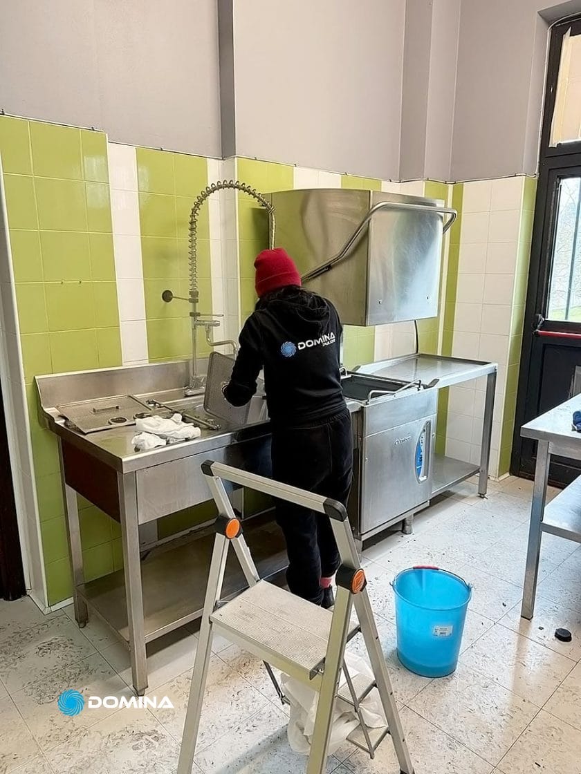 Una persona pulisce diligentemente un grande lavandino di metallo nella cucina di un ristorante, in piedi su una scala a pioli. Un secchio blu è appoggiato sul pavimento contro la parete piastrellata di verde, a mostrare l'impeccabile lavoro di Domina Pulizie.