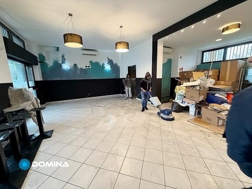 Le persone dell'Impresa di Pulizie stanno pulendo diligentemente una grande sala piastrellata al Ristorante Milano, con pile di scatole e sedie accatastate di lato. Le pareti sono parzialmente dipinte e le luci del soffitto sono appese sopra, assicurando un'atmosfera incontaminata per il servizio professionale.