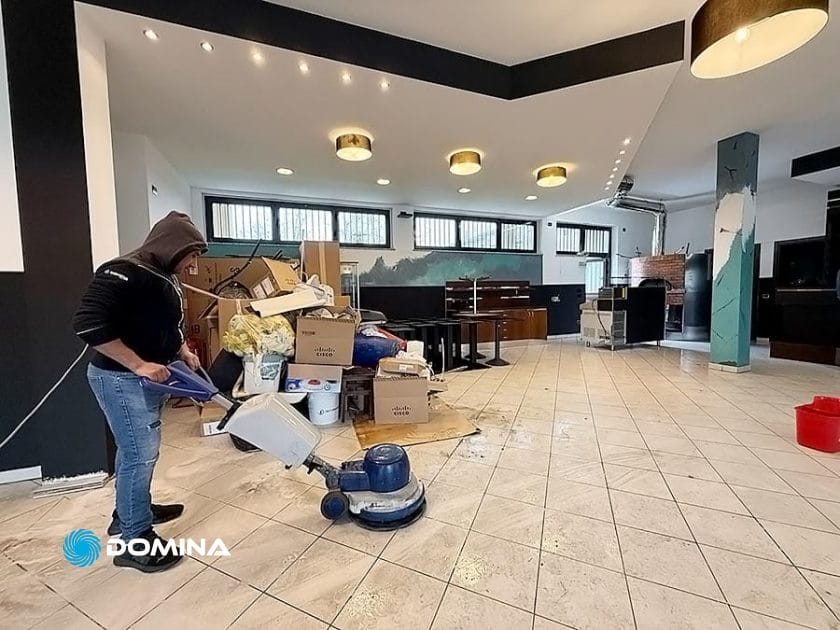 Un addetto dell'Impresa di Pulizie sta pulendo un pavimento piastrellato con una macchina in una stanza piena di scatoloni e detriti, assicurandosi che lo spazio sembri immacolato per il Ristorante Milano.