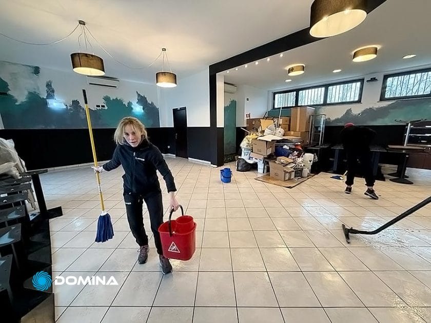 Nel cuore di Milano Centro, due professionisti eseguono meticolosamente le pulizie per ristorante. Uno trasporta uno straccio e un secchio rosso attraverso la grande stanza piastrellata piena di mobili e scatole accatastate, illuminata da luci a soffitto.