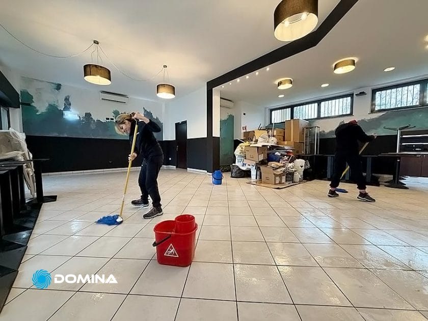 Due persone di un team di impresa di pulizie stanno pulendo diligentemente un pavimento piastrellato con stracci in una stanza ingombra di scatole e prodotti per la pulizia. Le luci del soffitto illuminano lo spazio, mentre le pareti parzialmente dipinte lasciano intuire lavori di ristrutturazione in corso.