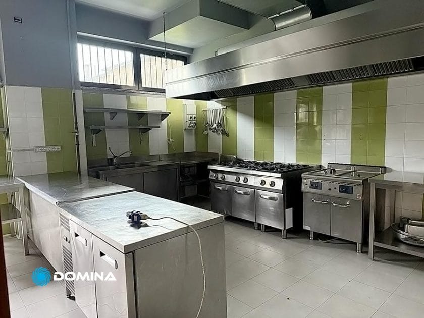 Questa cucina commerciale a Milano Centro è dotata di ripiani in acciaio inox, una grande cucina a gas e pareti piastrellate di verde e bianco. Con una cappa industriale e attrezzature da cucina visibili sugli scaffali, è lo spazio ideale per i servizi di Pulizie per Ristorante.