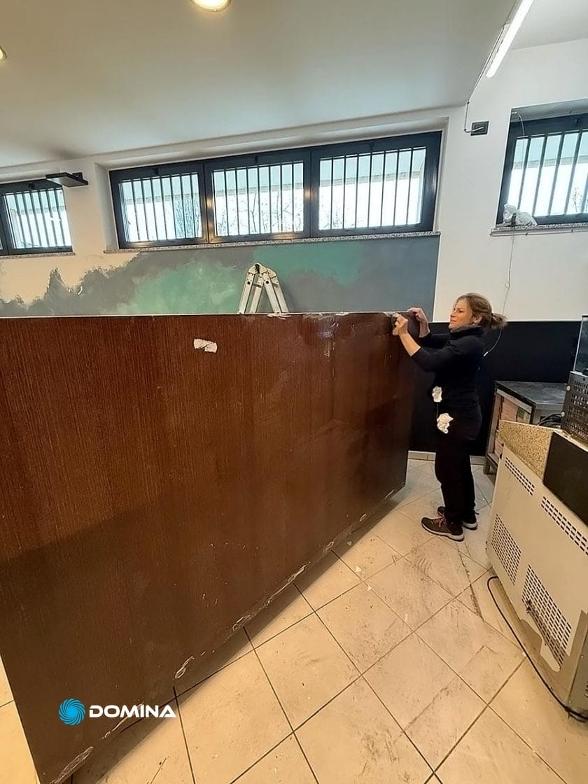 In una stanza con le finestre sbarrate, una persona afferra il bordo di un grande pannello di legno scuro. Nelle vicinanze, una scala suggerisce un lavoro in corso. La scena sembra quella di un riordino dietro le quinte al Ristorante Milano di Domina Pulizie.