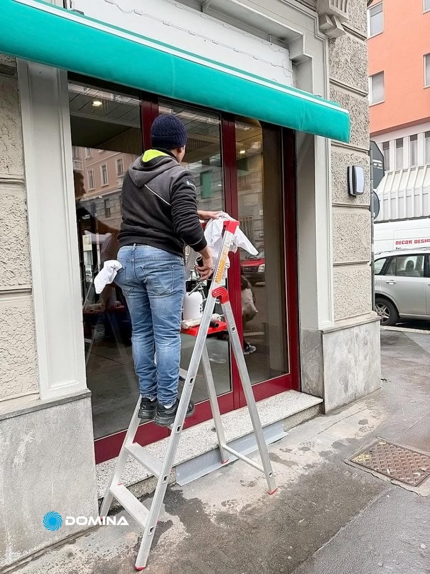 Un addetto di un'impresa di pulizie per ristoranti di Milano sta pulendo una porta a vetri mentre è in piedi su una scala a pioli all'esterno di un edificio con una tenda da sole verde acqua.