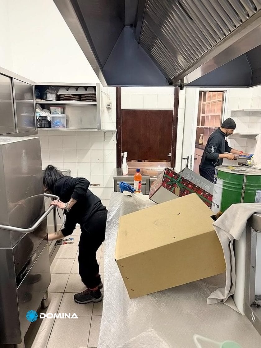 Due persone lavorano in modo efficiente in una cucina affollata. Una controlla la lavastoviglie mentre l'altra prepara il cibo al bancone. Una grande scatola di cartone e vari utensili da cucina li circondano, mostrando una scena che potrebbe trarre vantaggio dalla competenza di impresa pulizie per ristoranti Milano.